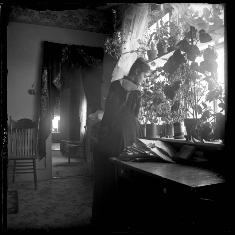 Kristin Holt | Historic Silver City, Idaho. Portrait of a woman, standing inside her home, with many potted houseplants. Image: Silver City, Owyhee County, Idaho, Public Domain. Image: P1960-139-22 Idaho State Archives.