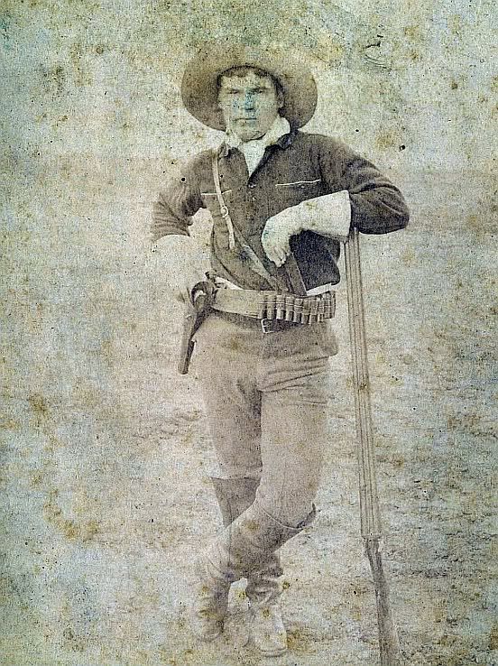 Kristin Holt | Armed Gunmen: Holsters, Braces, and Scabbards. Military Scout Junction City, (Fort Riley) Kansas ca 1890s. Photograph of armed military (Army) scout. Man standing and posing for photographer studio Ramsour & Pennel. He is wearing a wide brim hat, gauntlets, knee high boots, ammo belt with shot gun shells 
